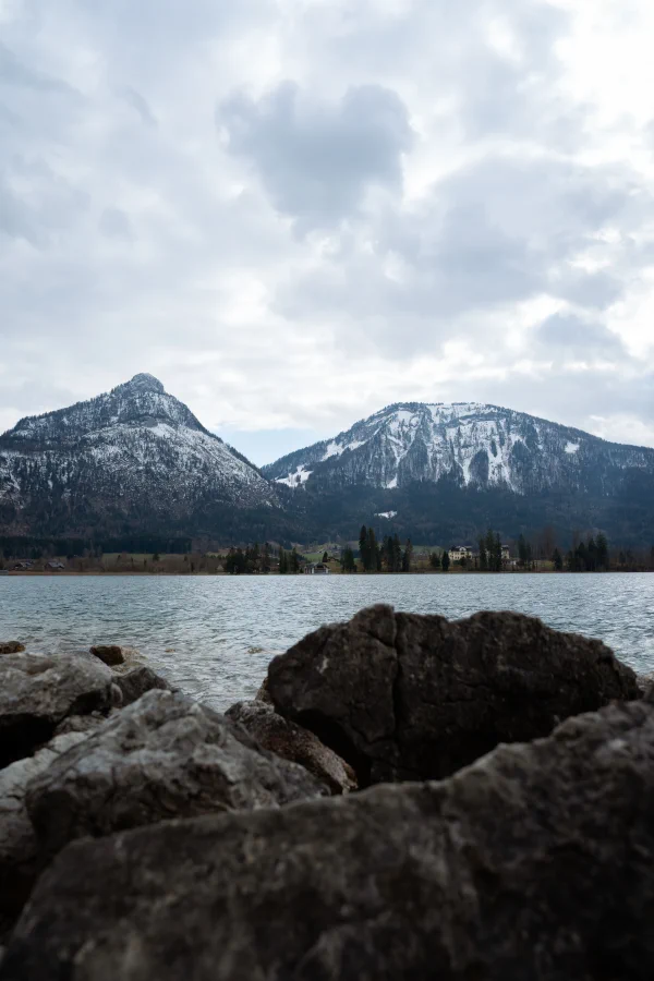 Rocks in the foreground, a still lake and snow-dusted peaks under an overcast sky