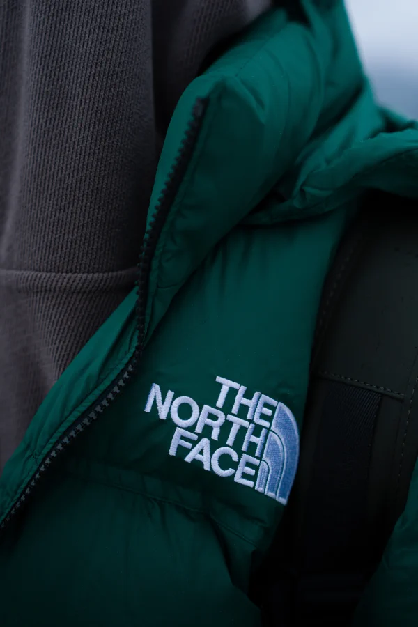Close-up of a green quilted hooded jacket with an embroidered brand logo and a backpack strap in mist