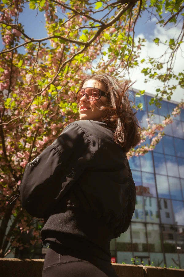 A portrait of a woman in a black jacket beneath a blossoming tree in front of a glass building