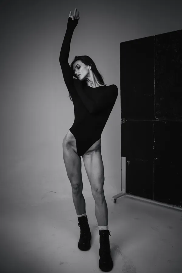A black-and-white studio portrait of a woman in black clothing with one arm raised