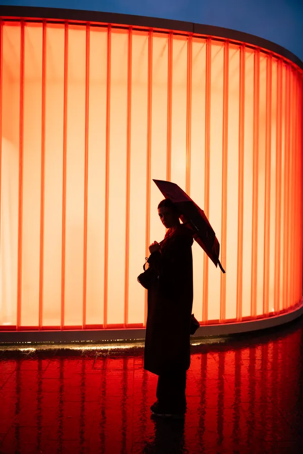 A person with an umbrella silhouetted against an orange-lit building